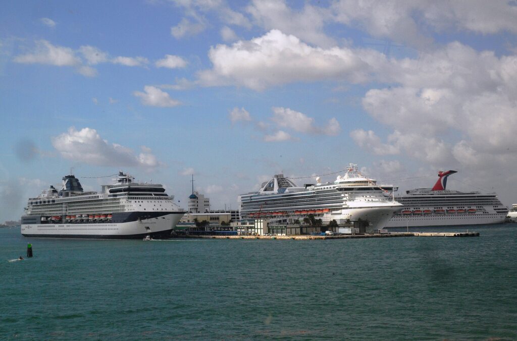 Port Everglades: tres cruceros blancos atracados en el muelle, uno con chimenea roja y azul en forma de cola, sobre agua verde azulada y cielo con nubes