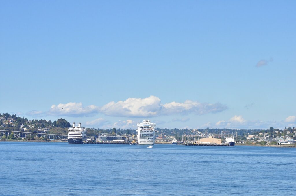 Port of Seattle: dos cruceros blancos atracados en la terminal de Smith Cove, vistos desde el agua azul de la bahía, con colinas arboladas detrás