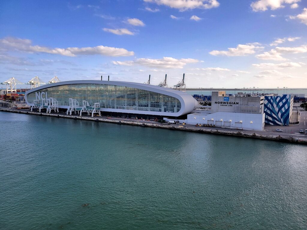 Terminal de cruceros de Miami: edificio blanco y curvo de vidrio con el letrero NCL junto al agua verde del puerto, con grúas al fondo y cielo azul