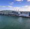 Terminal de cruceros de Miami: edificio blanco y curvo de vidrio con el letrero NCL junto al agua verde del puerto, con grúas al fondo y cielo azul