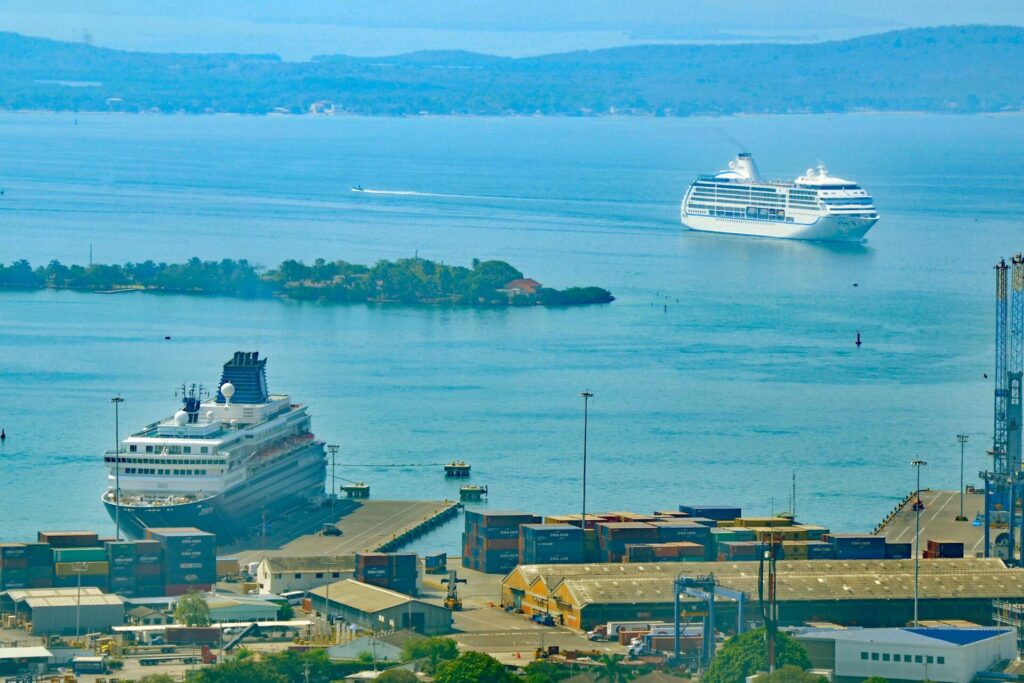 Terminal de cruceros de Cartagena: un crucero blanco atracado junto a contenedores del puerto mientras otro navega por la bahía azul, con una isla verde al fondo