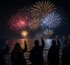 Pasajeros de espaldas brindando en la cubierta de un crucero mientras ven fuegos artificiales sobre el mar